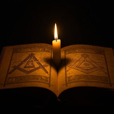 Livro aberto iluminado por uma vela, com esquadro e compasso nas páginas